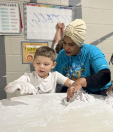 Young boy sitting at a table while an adult camp counselor assists him in an activity.